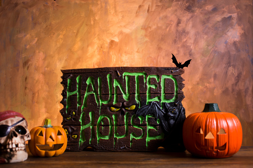 Bright orange background appears as to be an inferno with Halloween decorations in foreground.  Haunted House Sign and pumpkins.