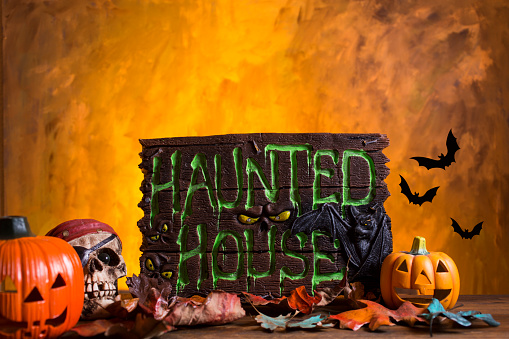 Bright orange background appears as to be an inferno with Halloween decorations in foreground.  Haunted House Sign and pumpkins.