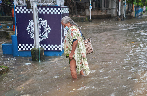 Deshapriya Park, Kolkata, 09/20/2021: An aged woman is seen wading through waterlogged street, before stepping in pavement. Due to fear of coronavirus pandemic, she is wearing protective face mask.