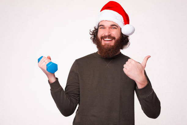 Happy young man, wearing Christmas hat is holding a little dumbbell while showing thumb up. stock photo