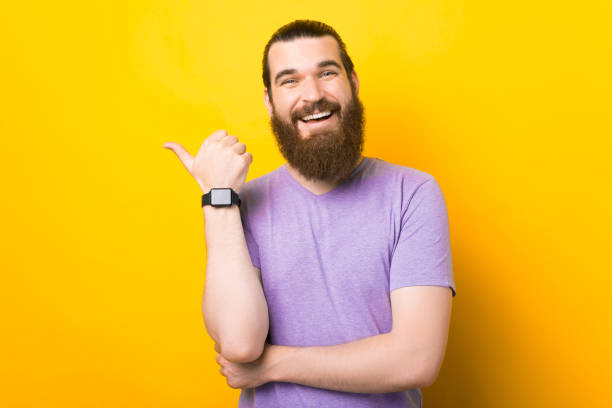 istock Bearded man is indicating with his thumb aside on the yellow background. 1341501331