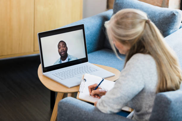 While teleconferencing, counselor takes notes as patient shares Sitting in her office, the unrecognizable female therapist takes notes as she video conferences with a mid adult male client. melancholy computer support specialist stock pictures, royalty-free photos & images