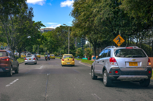 Bogotá, Colombia - September 18, 2021: The Driver's point of view on Carrera Septima in the Cedritos area of the  capital city of Bogota in the South American country of Colombia. The traffic is seen approaching the Calle 134 intersection. On either side of the road are mainly residential apartments and a few office buildings. Healthy trees grow everywhere in the capital City. It is a sunny morning. The elevation at street level is 8,660 feet above mean sea level. Horizontal format. Copy space.