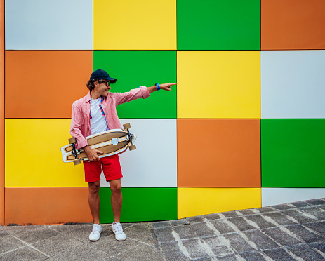 a middle-aged man, holding a skateboard while pointing his finger. the wall background is colorful.