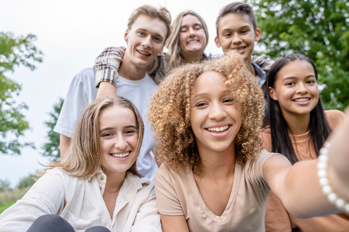 A group of ethnically diverse high school students stand closely together posing for a photo.  They are each dressed casually in hues of peach and pink as they smile.  The focus is on the a curly haired girl in front who has her hand outstretched holding her cell phone as she takes a selfie of the group.