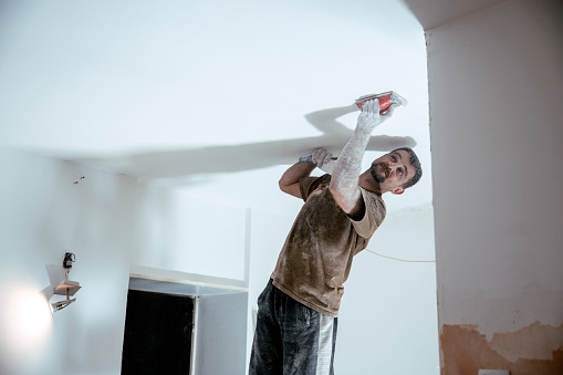 Man using sand paper preparing ceiling for the paint, standing on the ladders in dusty apartment