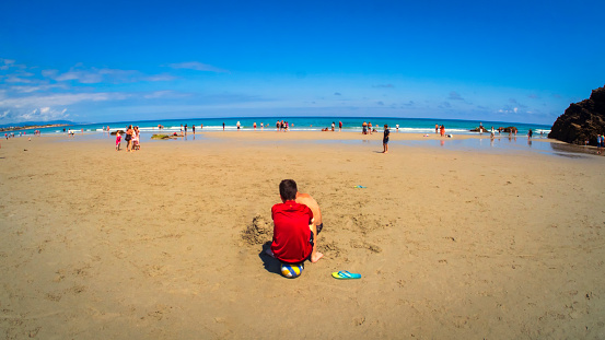 Ribadeo, Galicia, Spain. 07/26/2021 A boy in a red T-shirt plays with the sand on the beach of Las Catedrales on a blue-sky summer day.