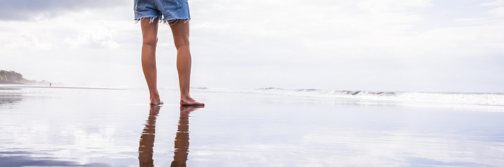 Panoramic photo of the legs of a woman standing on a beach with her silhouette and the sky reflected in the water.