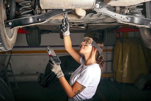 Young latina woman wearing goggles repairs a car with a wrench.