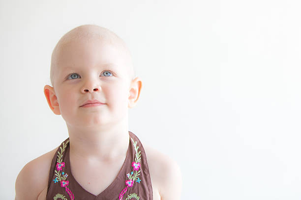 Young child with cancer in brown dress in front of a white background. Her blue eyes are looking upwards. Landscape composition leaves white space on the right side.