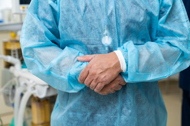 poważny lekarz w fartuchu ochronnym przygotowuje pacjenta operuje w nowoczesnej klinice. chirurg płci męskiej stojący ze skrzyżowanymi ramionami przy swoim gabinecie - examination gown zdjęcia i obrazy z banku zdjęć