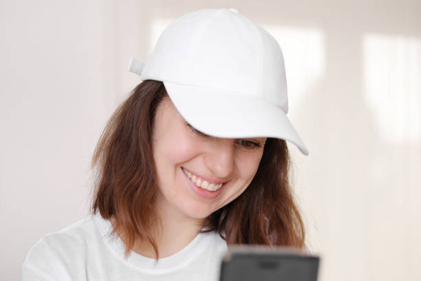 Female model wearing a white baseball cap. White cap mockup, template for picture, text or logo. Girl with a cap holding smarthphone. Free space, copy space. Female model wearing a white baseball cap. White cap mockup, template for picture, text or logo. Girl with a cap holding smarthphone. Free space, copy space woman wearing baseball cap stock pictures, royalty-free photos & images