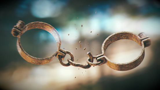 Rusty metallic handcuffs with a broken chain link and pieces of metal falling off, on a defocused bright background.