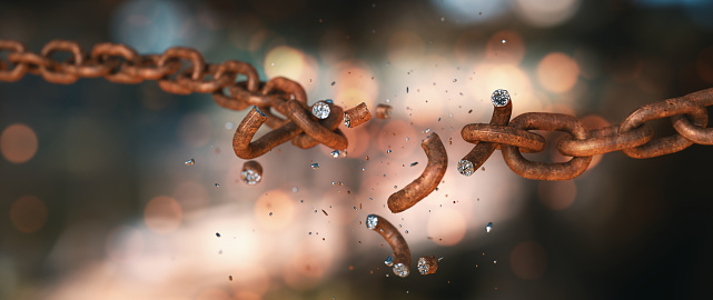 Extreme close-up view on a rusty old chain in the process of breaking into many pieces flying off, isolated on a defocused background with lens glare. Wide horizontal composition.