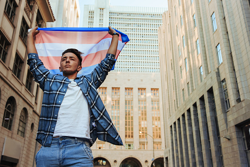 Nonconforming man raising the transgender flag outdoors. Confident young transgender man celebrating gay pride in the city. Young gender nonconforming man standing with the pride flag.
