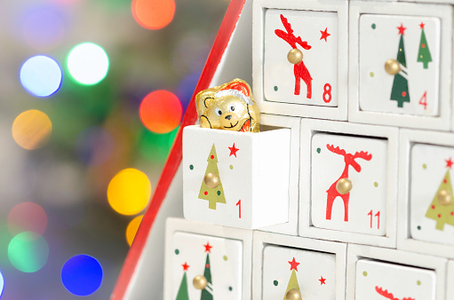 Close-up of the first compartment of a wooden advent calendar showing a bear's head candy. Background with defocused light on christmas tree.