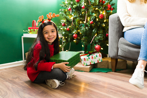 I feel so happy. Adorable latin girl sitting at home with her mom and opening presents during Christmas eve