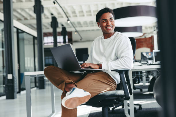 Cheerful software developer smiling in an office Cheerful software developer smiling in an office. Happy young businessman looking away while working on a laptop in a modern workplace. Creative businessman working on a new project. creative space photos stock pictures, royalty-free photos & images