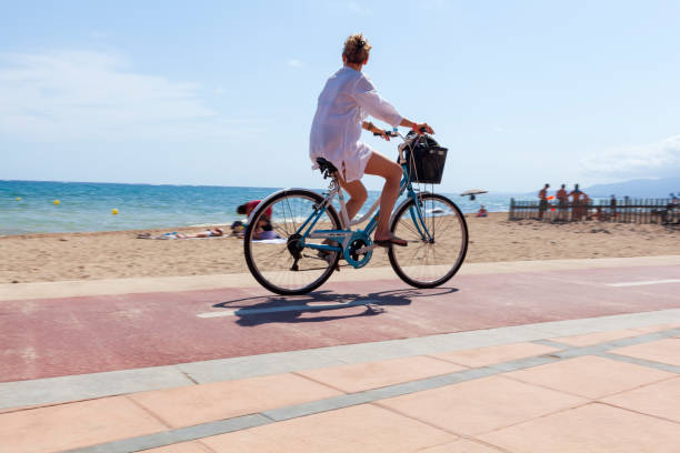 young woman riding bike along the shoreline of the beach of Cambrils, Spain. Sunny summer day. Spain. August 03, 2021. Cambrils: young woman riding bike along the shoreline of the beach. Sunny summer day. cambrils stock pictures, royalty-free photos & images
