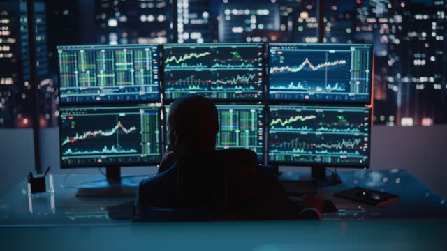 Hedge Fund Trader Working on a Computer with Multi-Monitor Workstation with Real-Time Stocks, Commodities and Exchange Market Charts. Businessman Works in Investment Bank Downtown Office at Night.