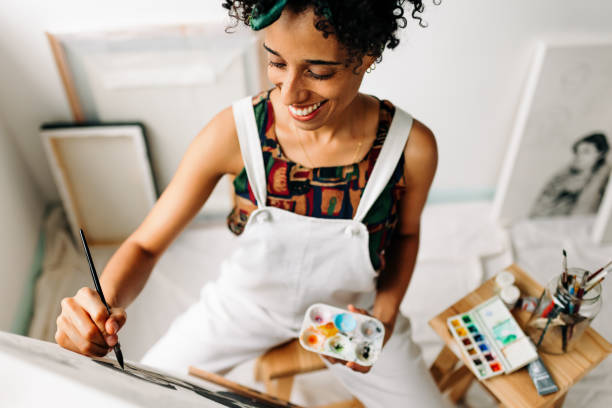 Happy female painter working in a workshop Happy young female painter working in a workshop. Young female painter smiling cheerfully while drawing on a canvas in her art studio. Creative young woman working on a new project. creative space photos stock pictures, royalty-free photos & images