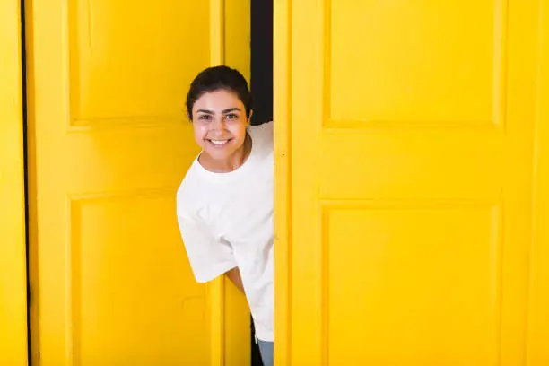 Young smiling indian woman peeking through yellow open door