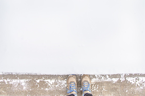 Person in brown sneakers standing outside on a rough and grunge concrete pavement at the edge of the white snow in winter season. Abstract minimal contrast concept idea with copy space.