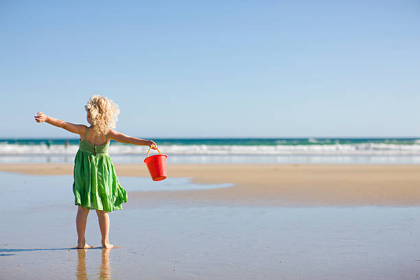 Little girl in green dress holding red bucket at the beach stock photo