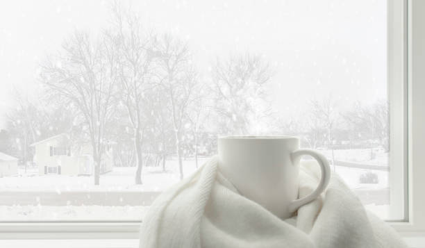 White winter concept of hot drink and winter accesseries Hot tea mug covered by white comfort soft wool scarf in front of frosted glass window in warm and cozy living room with lonely but beautiful landscape view snow falling outside. White winter concept. blizzard stock pictures, royalty-free photos & images