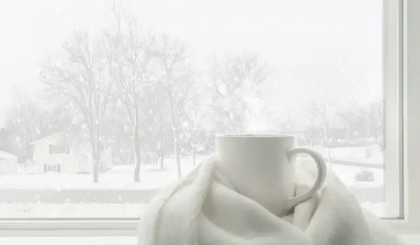 Hot tea mug covered by white comfort soft wool scarf in front of frosted glass window in warm and cozy living room with lonely but beautiful landscape view snow falling outside. White winter concept.