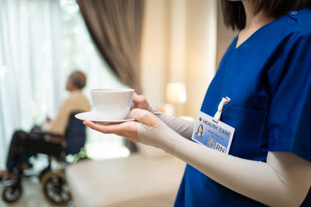 アジアの美しいセラピスト医師は、家の中で古い患者にコーヒーを提供しています。介護施設の若い女性看護師が車椅子で障害のある高齢者の世話をしています。医療保険サービスの概念 - senior adult nursing home eating home interior ストックフォトと画像