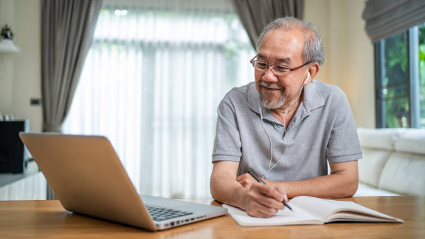ノートパソコンでオンラインで働くアジアの高齢者のビジネスマンのホームオフィス。成熟した年上の男性は、オンライン会議のビデオ通話で話し、covid-19パンデミック中に社内でコピーブ� - chinese ethnicity audio ストックフォトと画像