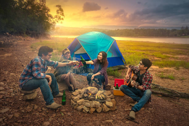 happy friends camping party playing music and enjoying bonfire in nature and lake, traveler camping on the mountain. with a relaxing drink in the sunset. add noise and film grain/soft focus filter. - bonfire beach fire barbecue imagens e fotografias de stock