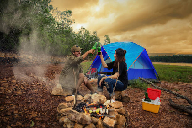 happy friends camping party and enjoying bonfire in nature and lake, traveler camping on the river/mountain. with a relaxing drink in the sunset. the concept of hiking/camping and relax. - bonfire beach fire barbecue imagens e fotografias de stock