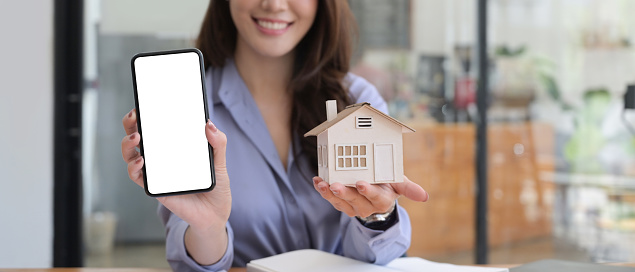 Smiling woman showing smart phone with blank screen and holding house model.