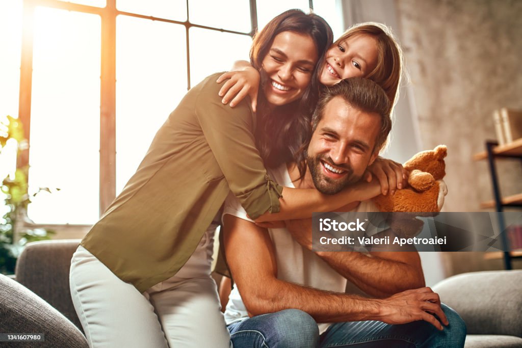 Family at home Happy dad and mom with their cute daughter and teddy bear hug and have fun sitting on the sofa in the living room at home. Family Stock Photo