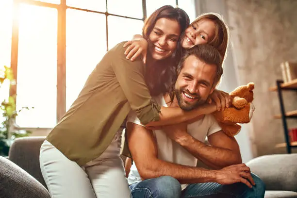 Happy dad and mom with their cute daughter and teddy bear hug and have fun sitting on the sofa in the living room at home.