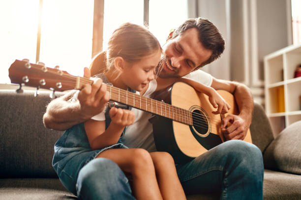 Family at home Happy dad teaches his cute daughter to play the guitar while sitting on the sofa in the living room at home. Happy Father's Day. family at home stock pictures, royalty-free photos & images