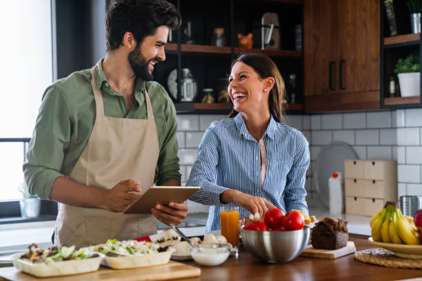 Beautiful young couple is using a digital tablet and smiling while cooking in kitchen at home stock photo