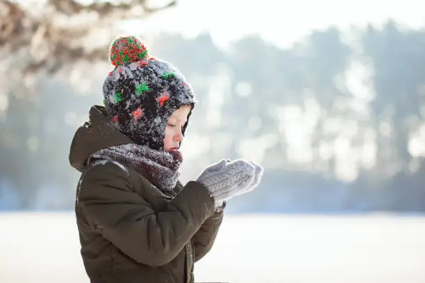 Portrait of a cute little boy in warm clothes blowing on snow outdoors during snowfall in winter sunny day