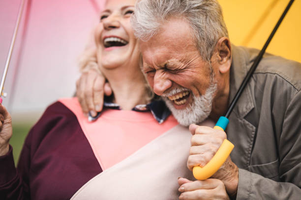 cheerful mature couple having fun while laughing under umbrellas. - umbrella senior adult couple autumn imagens e fotografias de stock