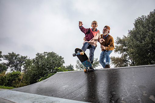 Low angle view of happy senior woman riding skateboard on a ramp while her husband is assisting her in a skate park. Copy space.