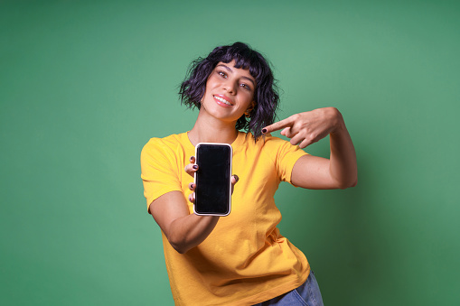 Cheerful happy brunette woman in t-shirt showing blank smartphone screen and pointing on it while looking at the camera smiling over green background