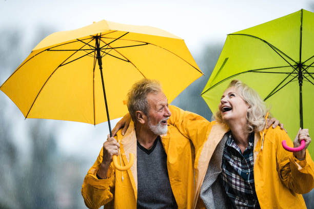 couple de personnes âgées heureux en imperméable parlant sous des parapluies un jour de pluie. - umbrella senior adult couple autumn photos et images de collection