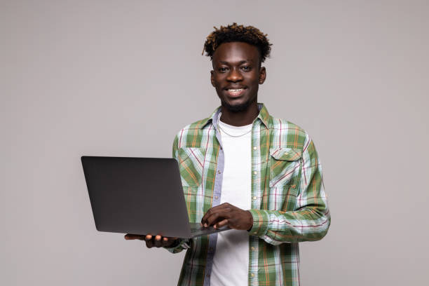 istock Young smiling african man standing and using laptop computer over gray background 1341621125