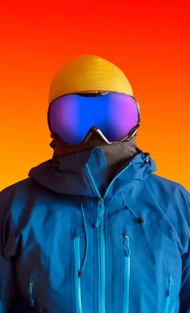Vertical portrait of a skier dressed in blue hardshell jacket and ski goggles placed over a vivid orange background.