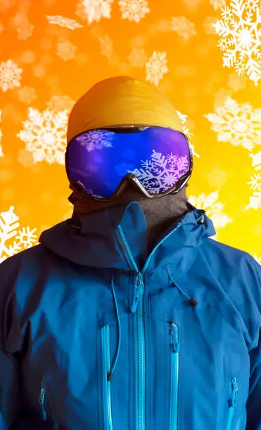 Vertical portrait of a skier dressed in blue hardshell jacket and ski goggles with falling snowflakes reflected in the glass. All placed over a vivid orange background with snow.