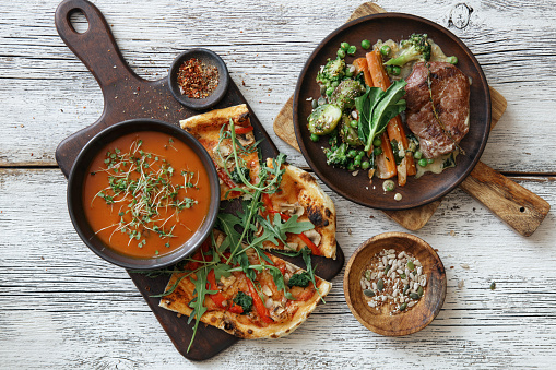 Steak with glazed carrot, green pea and Brussels sprouts. Pizza with vegetables and arugula. Roasted red bell pepper soup. Flat lay top-down composition on white wooden background.