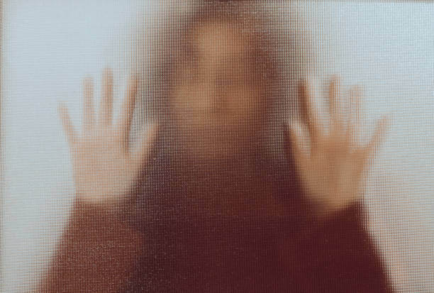Domestic abuse victim with hands pressed against glass window Back lit image of the silhouette of a woman with her hands pressed against a glass window. The silhouette is distorted, and the arms elongated, giving an alien-like quality. The image is sinister and foreboding, with an element of horror. It is as if the 'woman' is trying to escape from behind the glass. harassment stock pictures, royalty-free photos & images