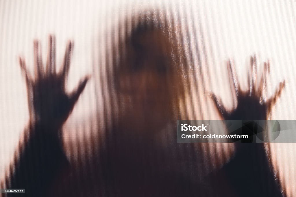 Domestic abuse victim with hands pressed against glass window Back lit image of the silhouette of a woman with her hands pressed against a glass window. The silhouette is distorted, and the arms elongated, giving an alien-like quality. The image is sinister and foreboding, with an element of horror. It is as if the 'woman' is trying to escape from behind the glass. Sexual Assault Stock Photo